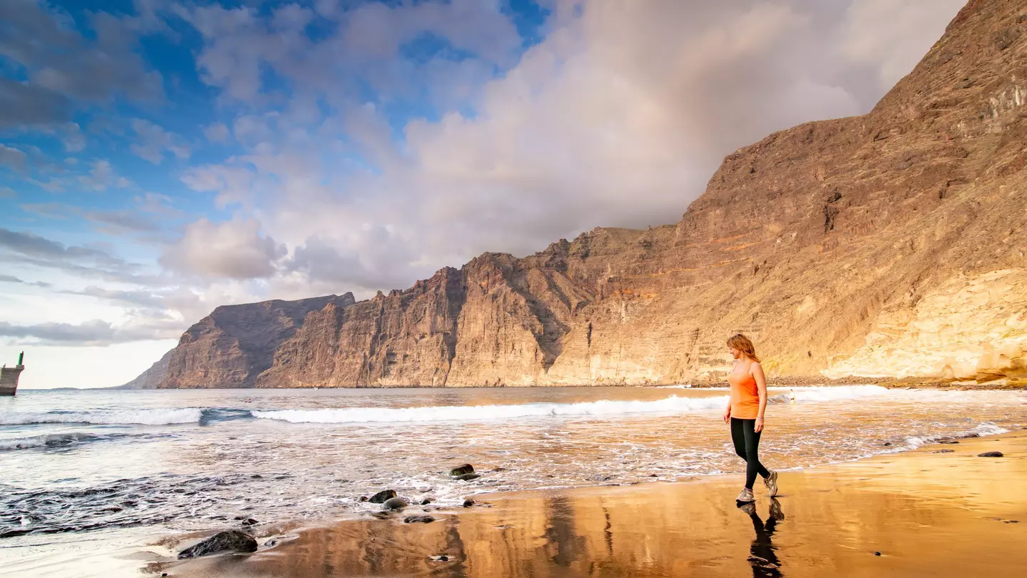 Adult woman walking along the beach in Tenerife, backed by huge sheer rocky cliffs