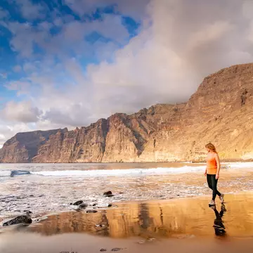 Adult woman walking along the beach in Tenerife, backed by huge sheer rocky cliffs