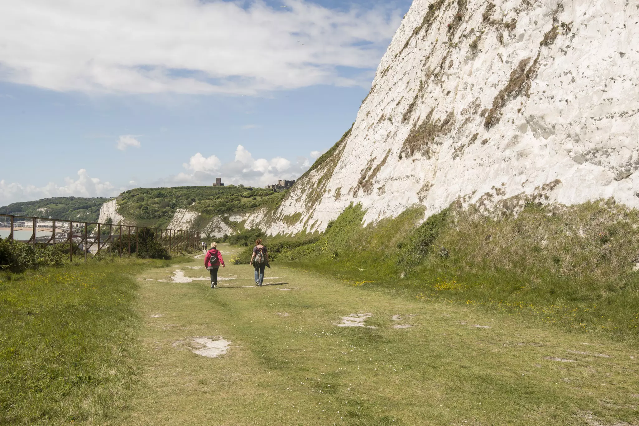Two people walking on a grass path by white cliffs toward a castle on a far hill.
