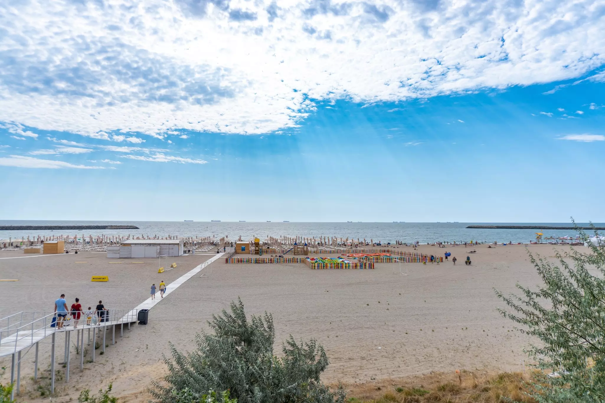 Wide sandy beach bisected by wooden boardwalk. Loungers and umbrellas fill the sand with the ocean in the distance and streams of sunlight come through thin, white clouds.
