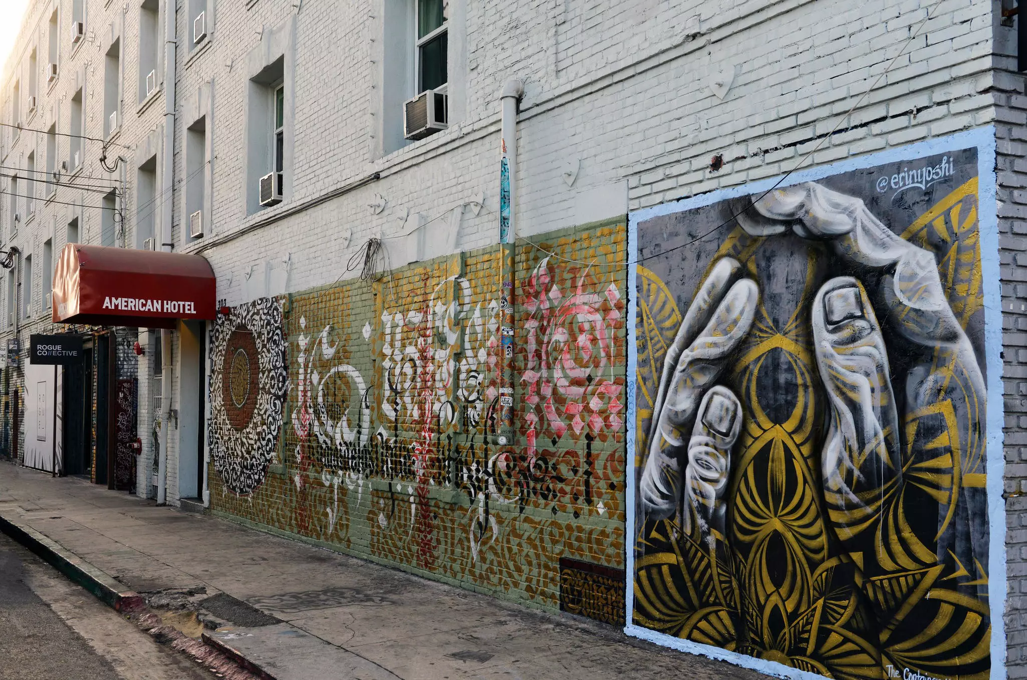 Wide shot of side of brick building with "American Hotel" written on a red awning and two pieces of street art, one with a variety of script letters and the other with two large hands holding an abstract botanical image.