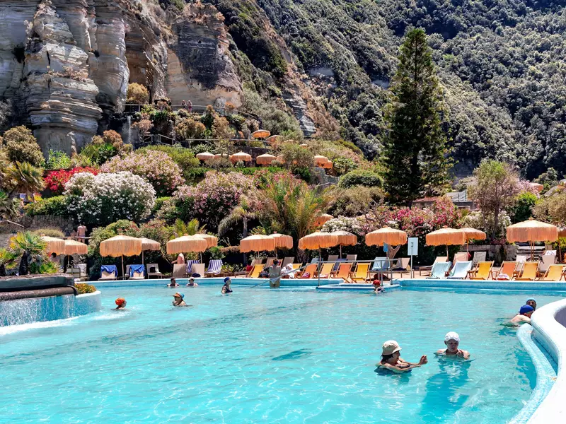 People swim in a thermal pool, with umbrellas and lounge chairs on the perimeter and mountains in the background. 