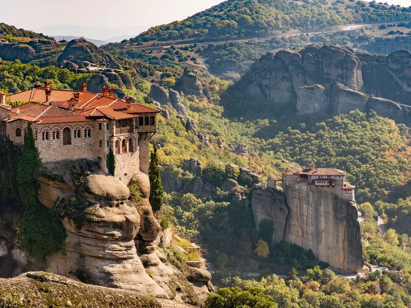 A red-roofed monstery sits high on a rocky outcrop in a lush mountain region. 