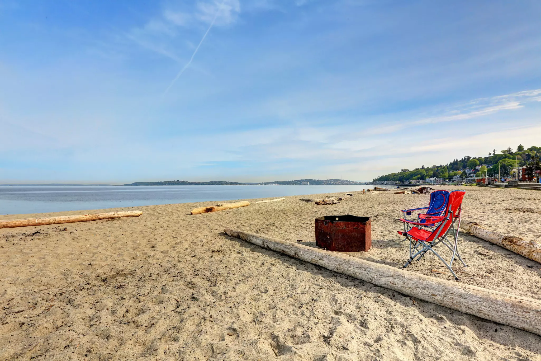 Two camping chairs on a beach in front of a fire pit near somme driftwood.
