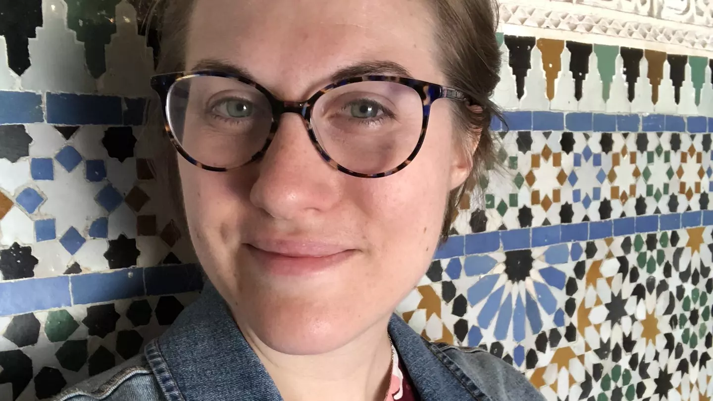 A woman wearing glasses looks into the camera in front of a tile wall.