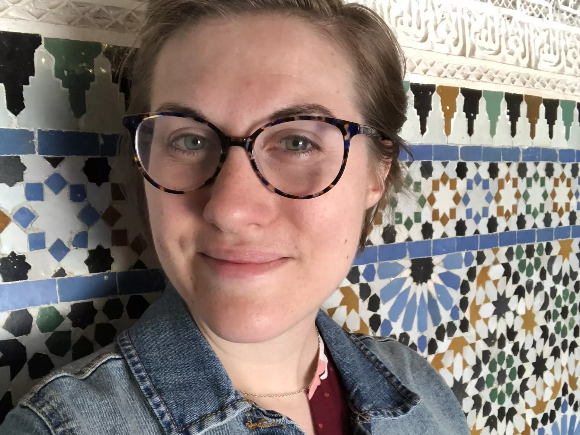 A woman wearing glasses looks into the camera in front of a tile wall.