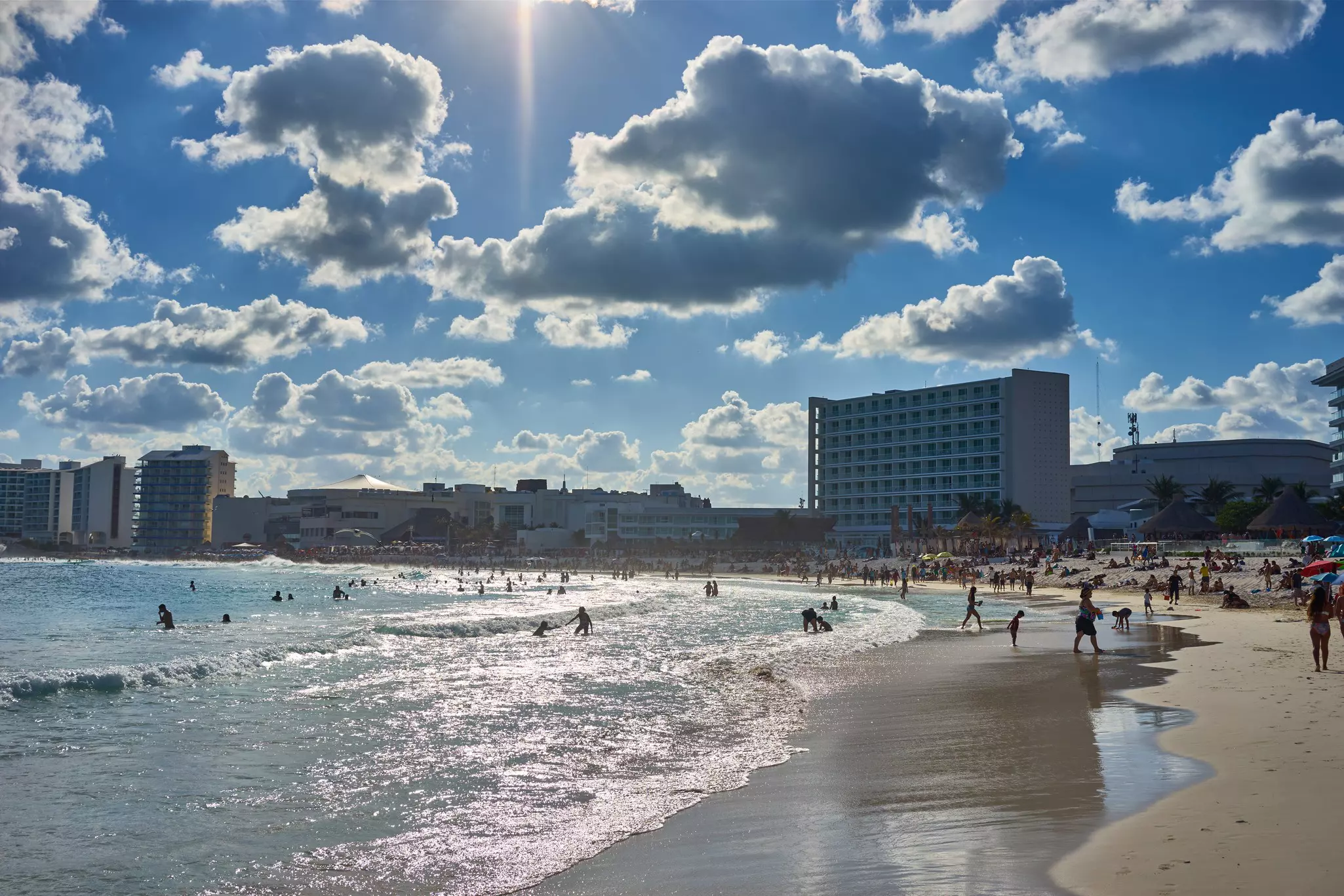 Gentle waves lap a beach, where people are seen walking on the sand, and swimming in the surf. Large hotels line the shore in the distance.