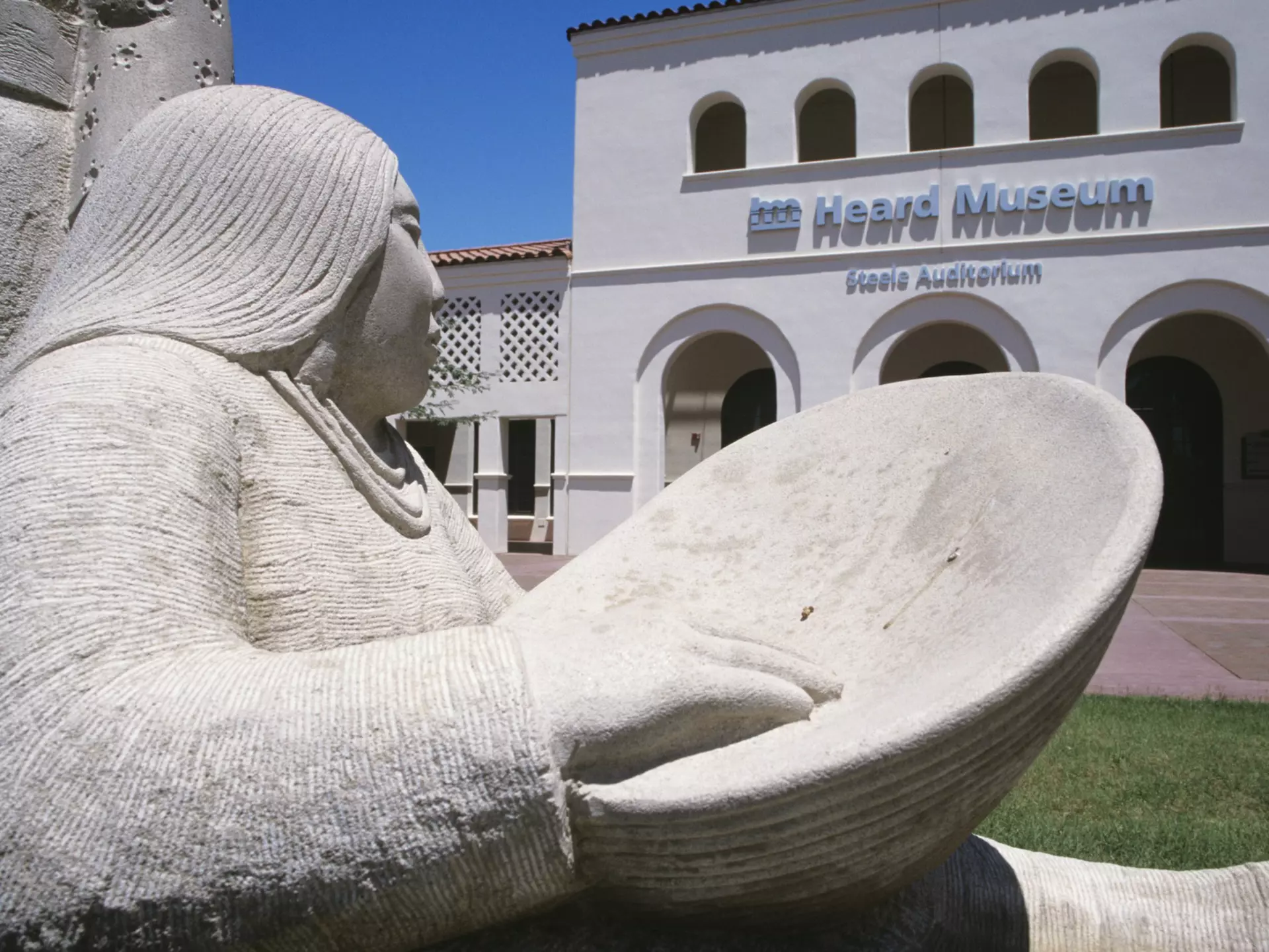 A sculpture of a Native American woman with a bowl sits outside the white-walled Heard Museum in Phoenix, Arizona, USA