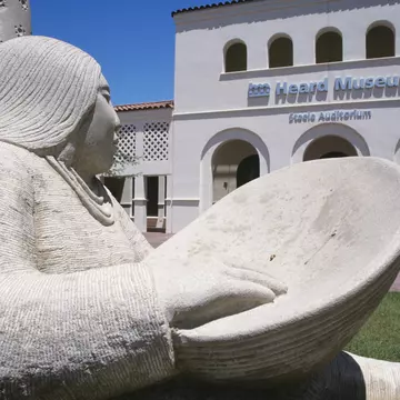 A sculpture of a Native American woman with a bowl sits outside the white-walled Heard Museum in Phoenix, Arizona, USA
