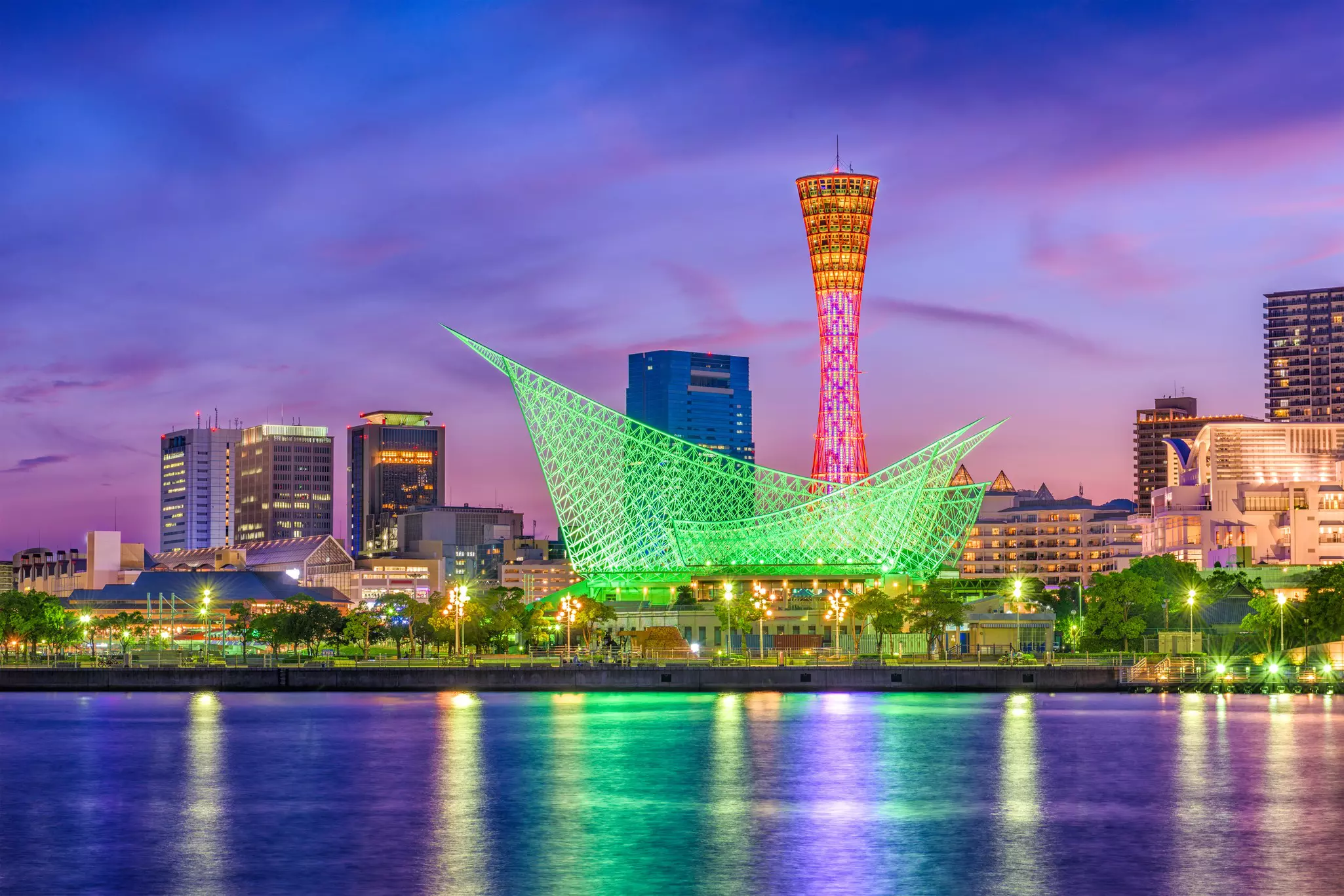 A city waterfront lit up at night with a tall latticed tower glowing red and a sail-like structure in green reflecting in the harbour water