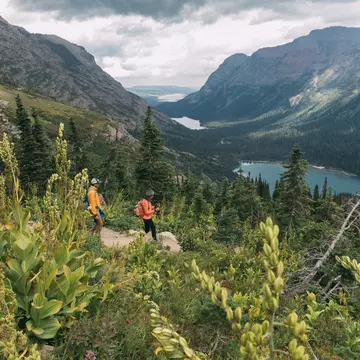 Two hikers follow a path through thick undergrowth downhill toward a lake