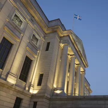 Exterior of a white stone museum with columns out front and a lighted lamppost in early evening.