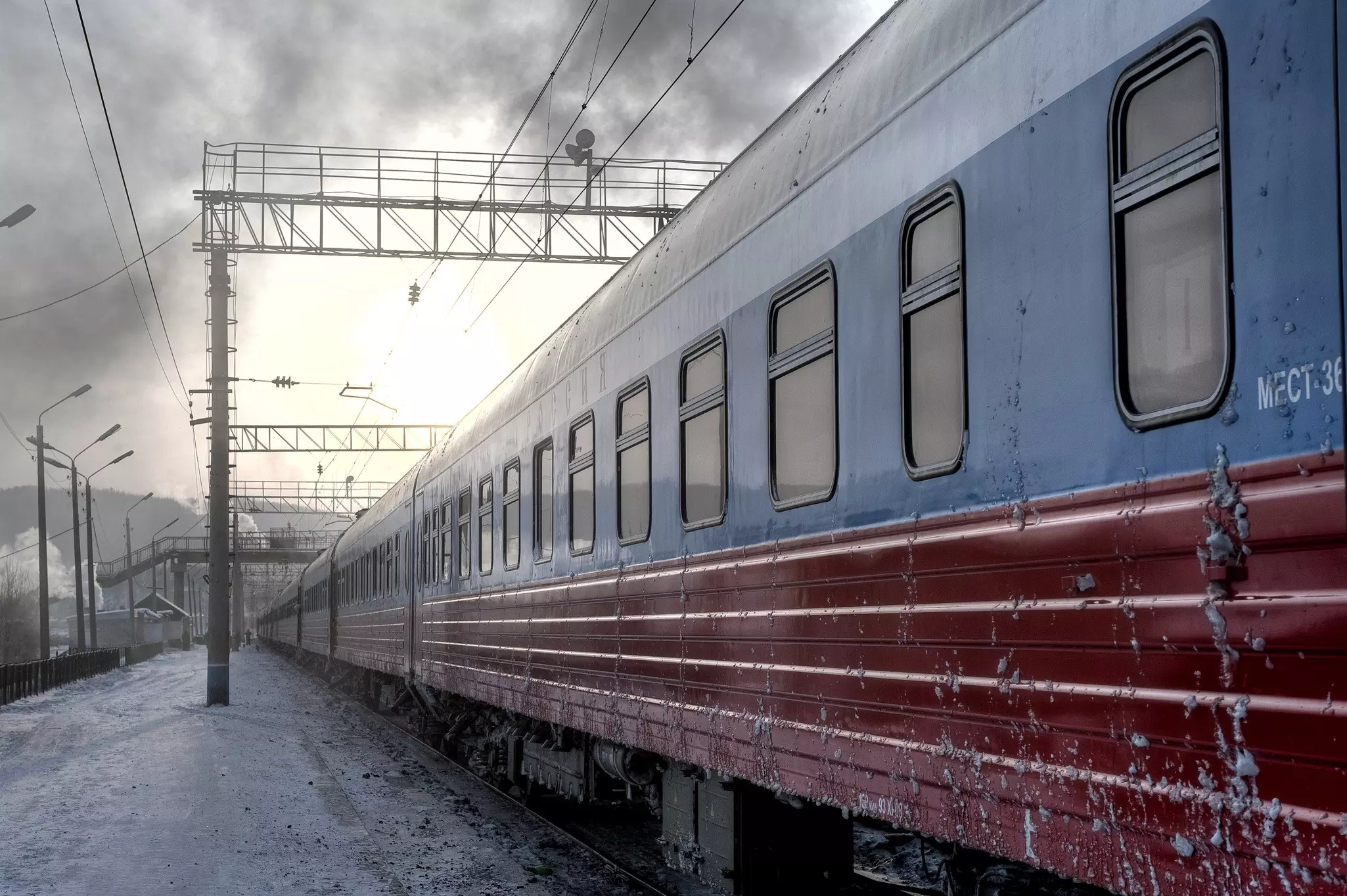 A red and grey train stand at an icy platform; grey clouds loom in the sky and there are many overhead power lines.
