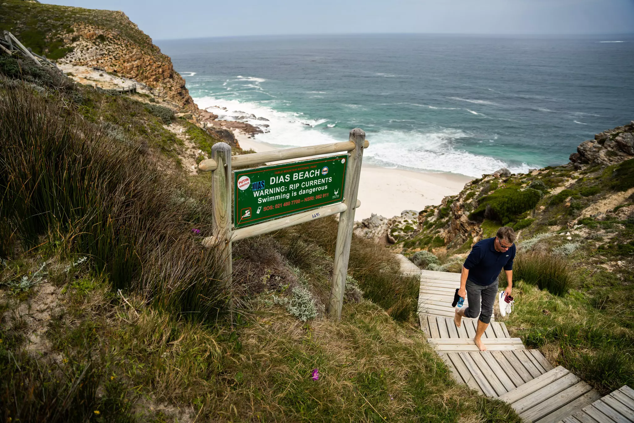 A barefoot man walks up wooden steps from a beach cove. Cliffs are visible on either side of the beach.