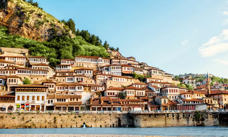 Tiers of white Ottoman houses with many windows stacked up a green hillside above a river, a minaret to the right