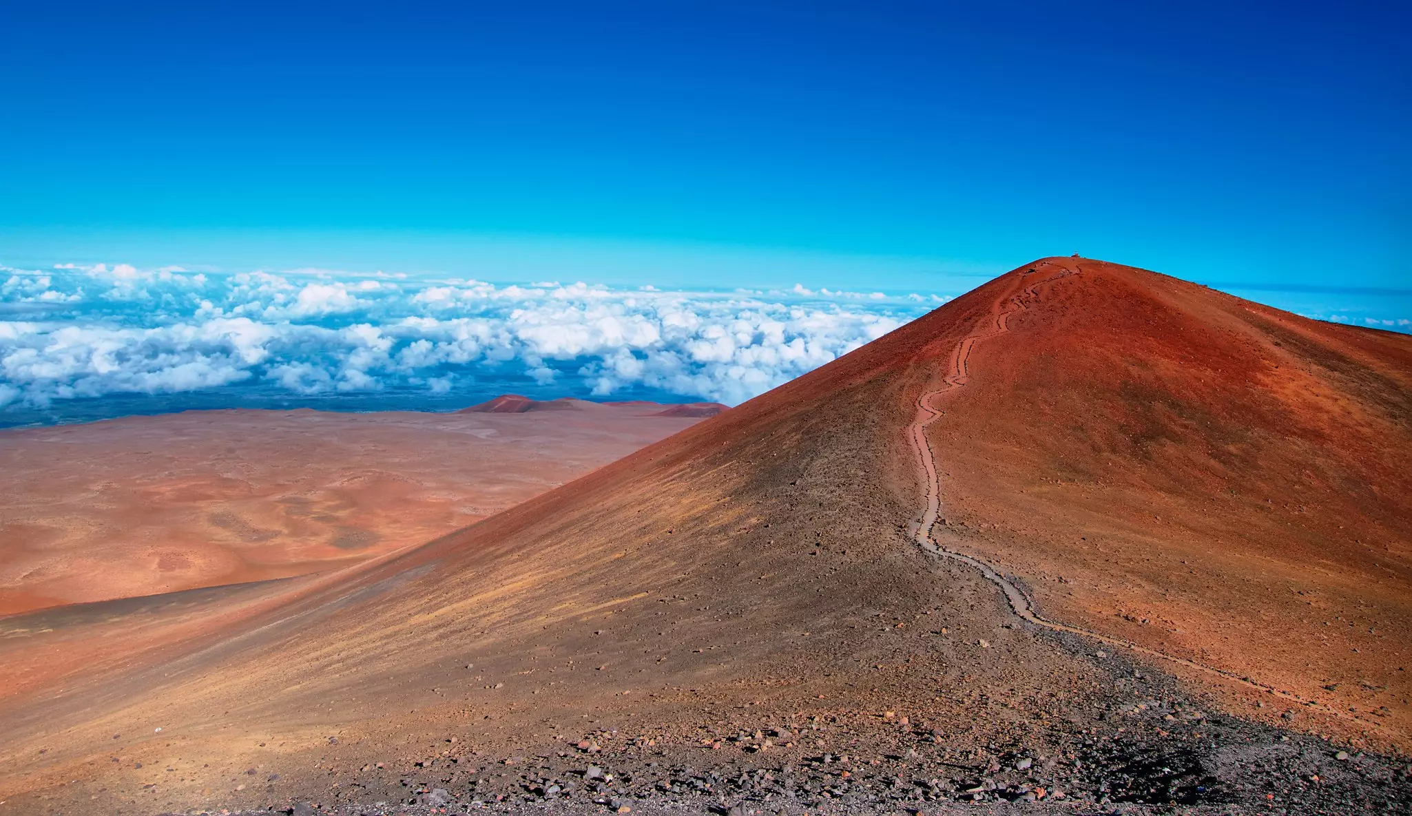 A narrow path leads to the summit of a red-dirt mountain with smaller hills and clouds in the distance and blue sky above.