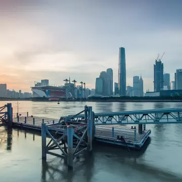 A metal pier and walkway on a wide river under an orange sunset sky, a skyline of tall towers across the water.