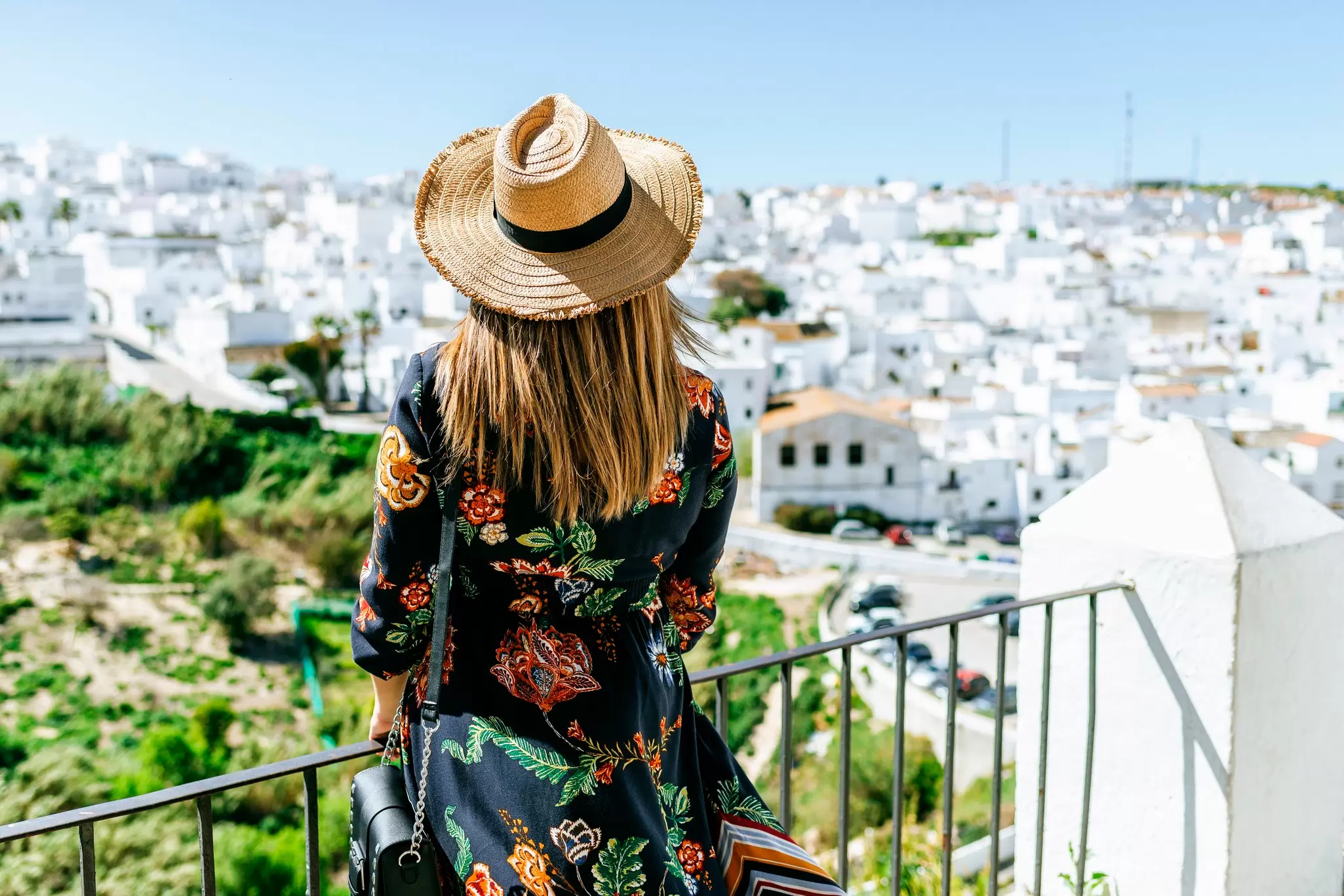 Woman in a floral dress and straw hat overlooking a hillside town of whitewashed houses from a railing