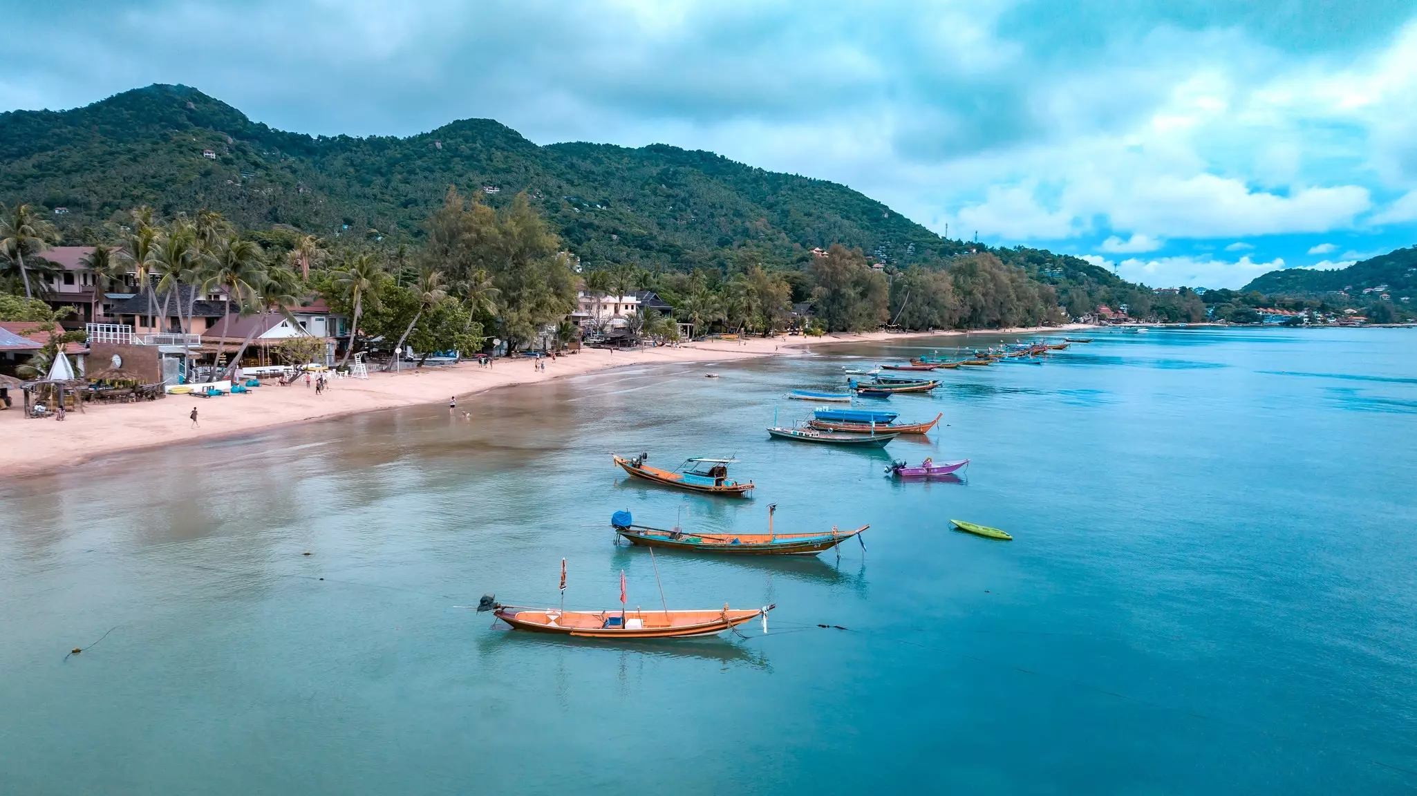 Traditional Thai longtail boats docked offshore near a sandy beach backed by palm trees.