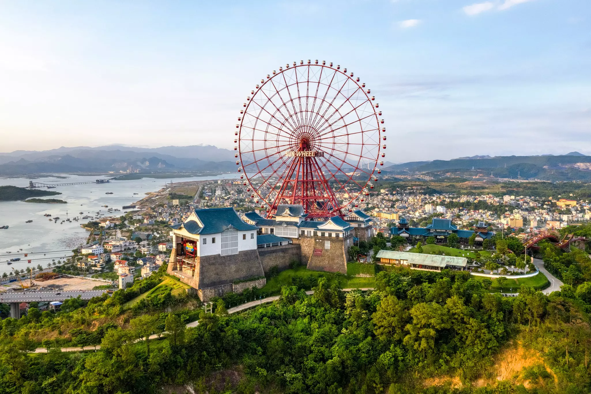 A large red Ferris wheel stands at a complex at the top of a hill overlooking a city and a bay.