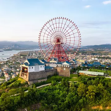 A large red Ferris wheel stands at a complex at the top of a hill overlooking a city and a bay.