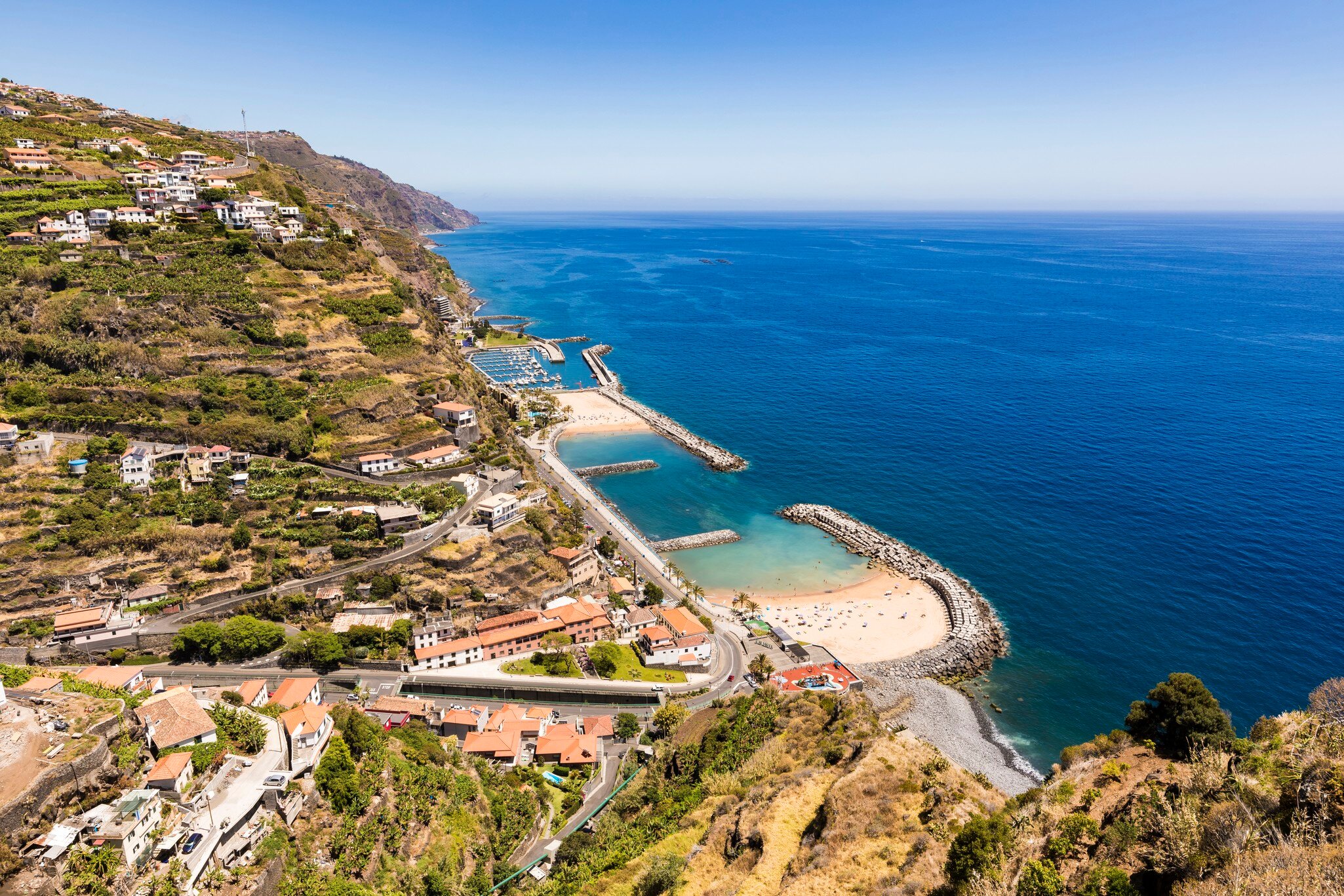 Hillside view over terraced slopes, red-roofed houses, a marina and two sandy coves protected by breakwaters