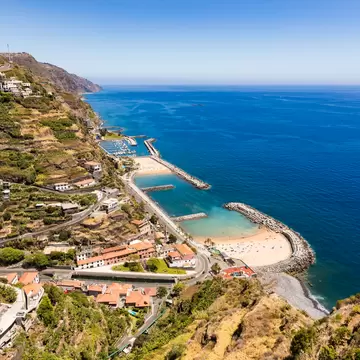 Hillside view over terraced slopes, red-roofed houses, a marina and two sandy coves protected by breakwaters