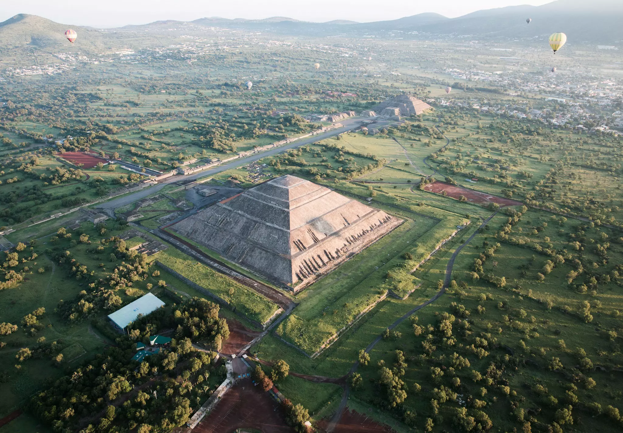 Aerial shot of pyramids, one in foreground and one in background, with a few hot air balloons in the sky and mountains in the distance.