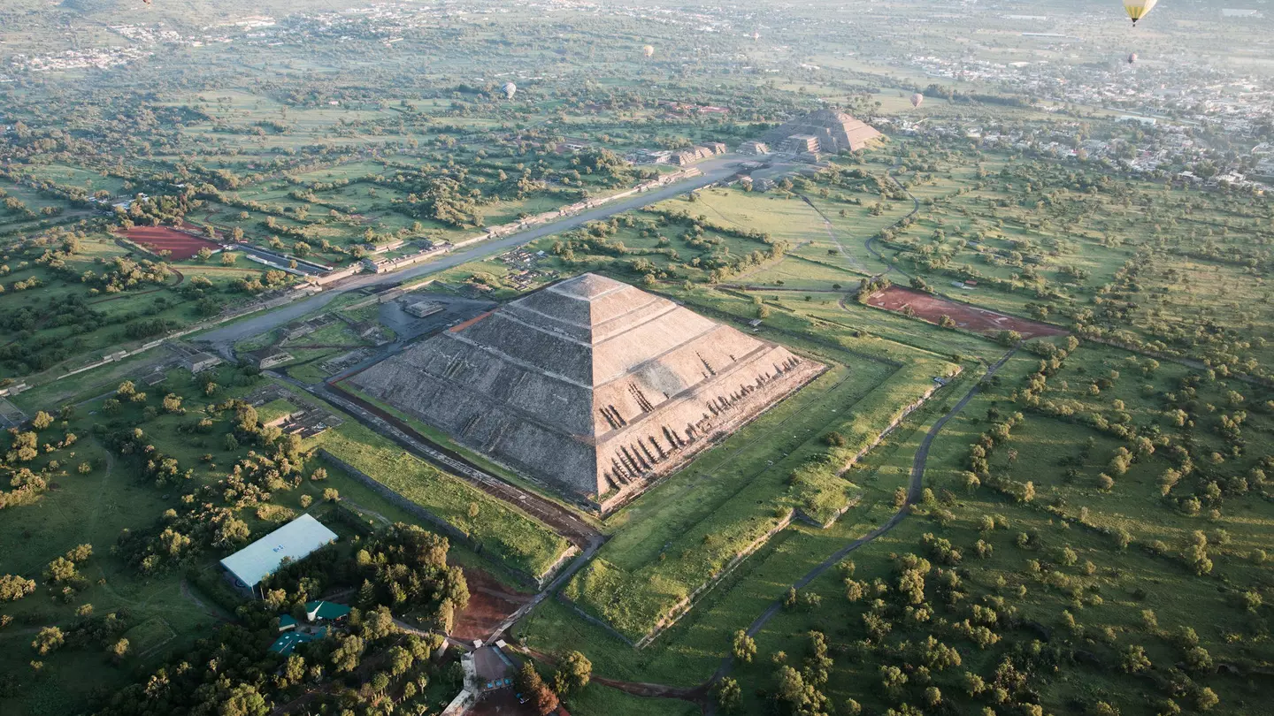 Aerial shot of pyramids, one in foreground and one in background, with a few hot air balloons in the sky and mountains in the distance.