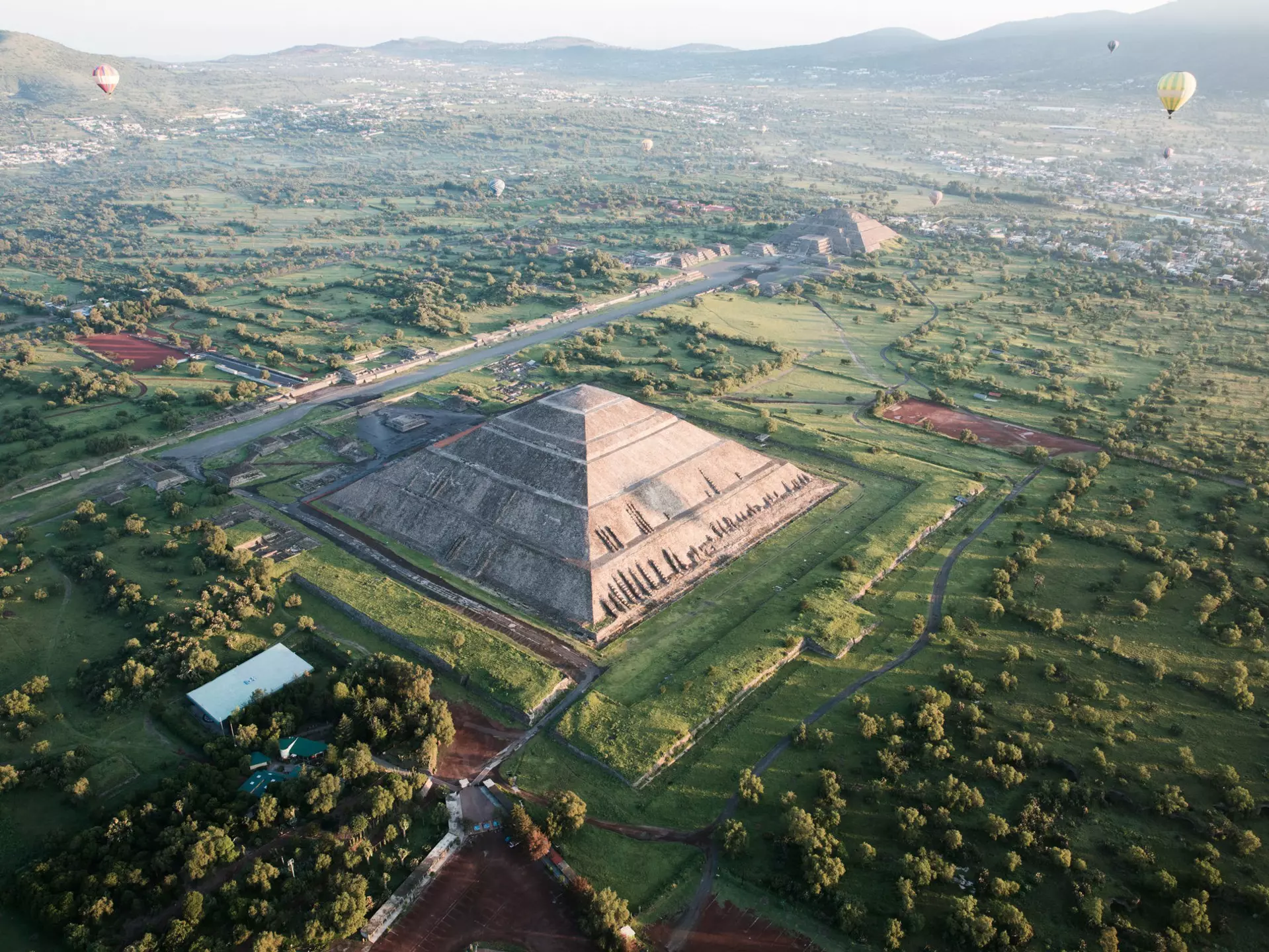 Aerial shot of pyramids, one in foreground and one in background, with a few hot air balloons in the sky and mountains in the distance.
