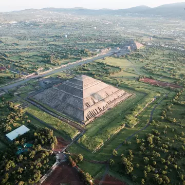 Aerial shot of pyramids, one in foreground and one in background, with a few hot air balloons in the sky and mountains in the distance.