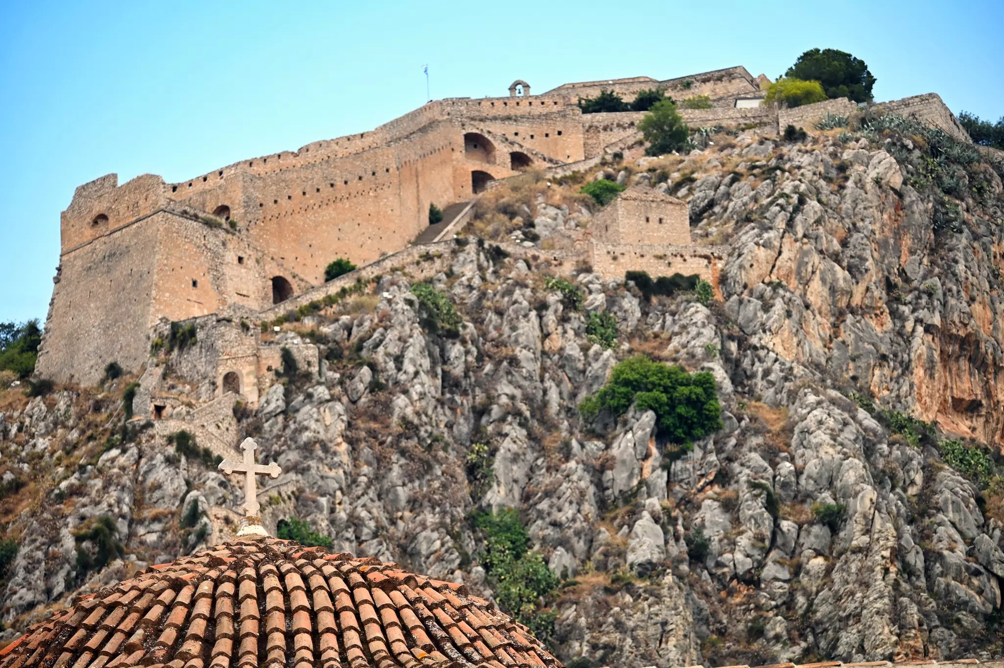 A cross on a tiled church dome is visible in the foreground in front of a mountaintop fortress, seen from below.