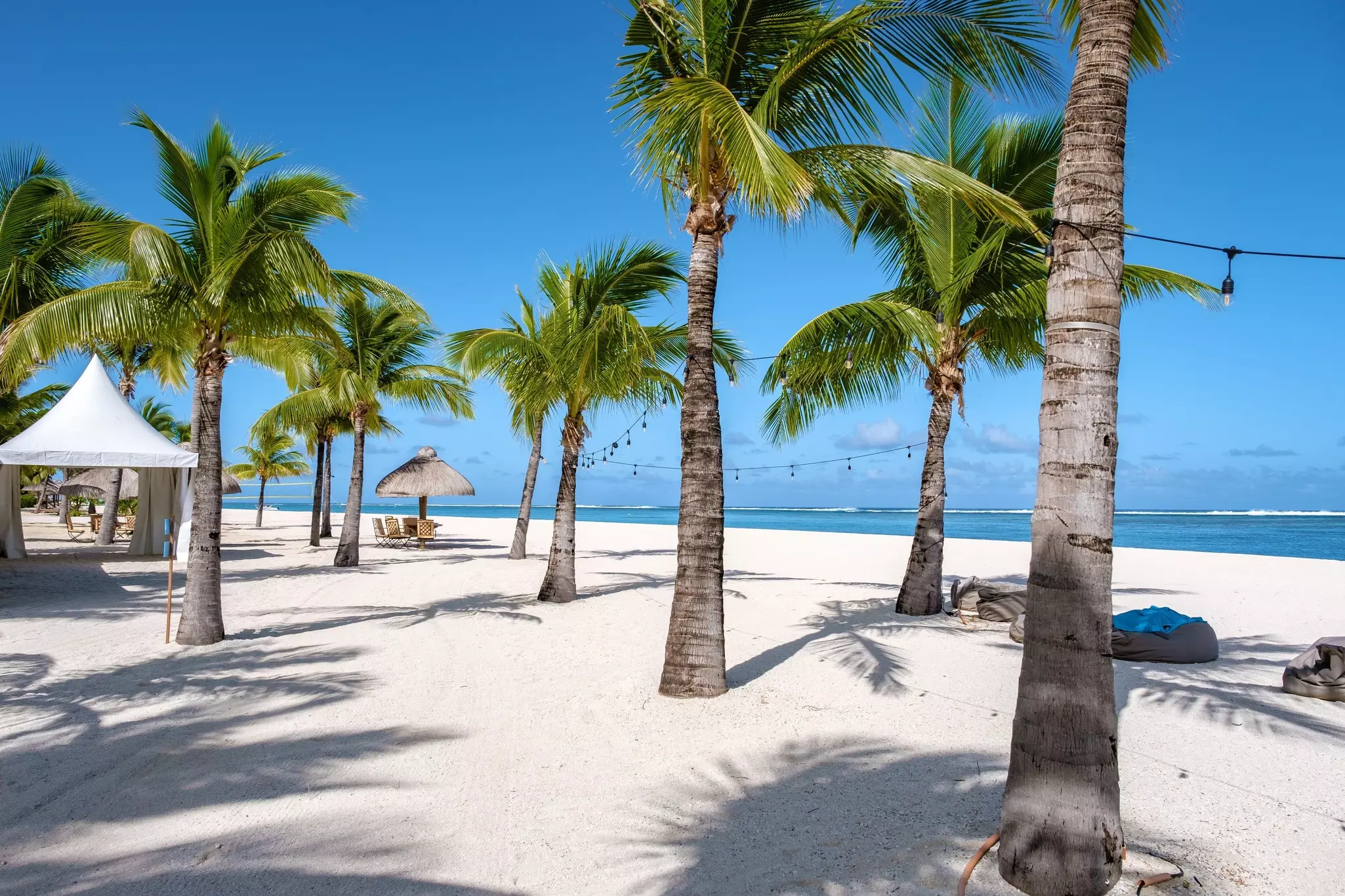 Pam trees and shelters offer some shade on a white-sand beach backed by a resort.