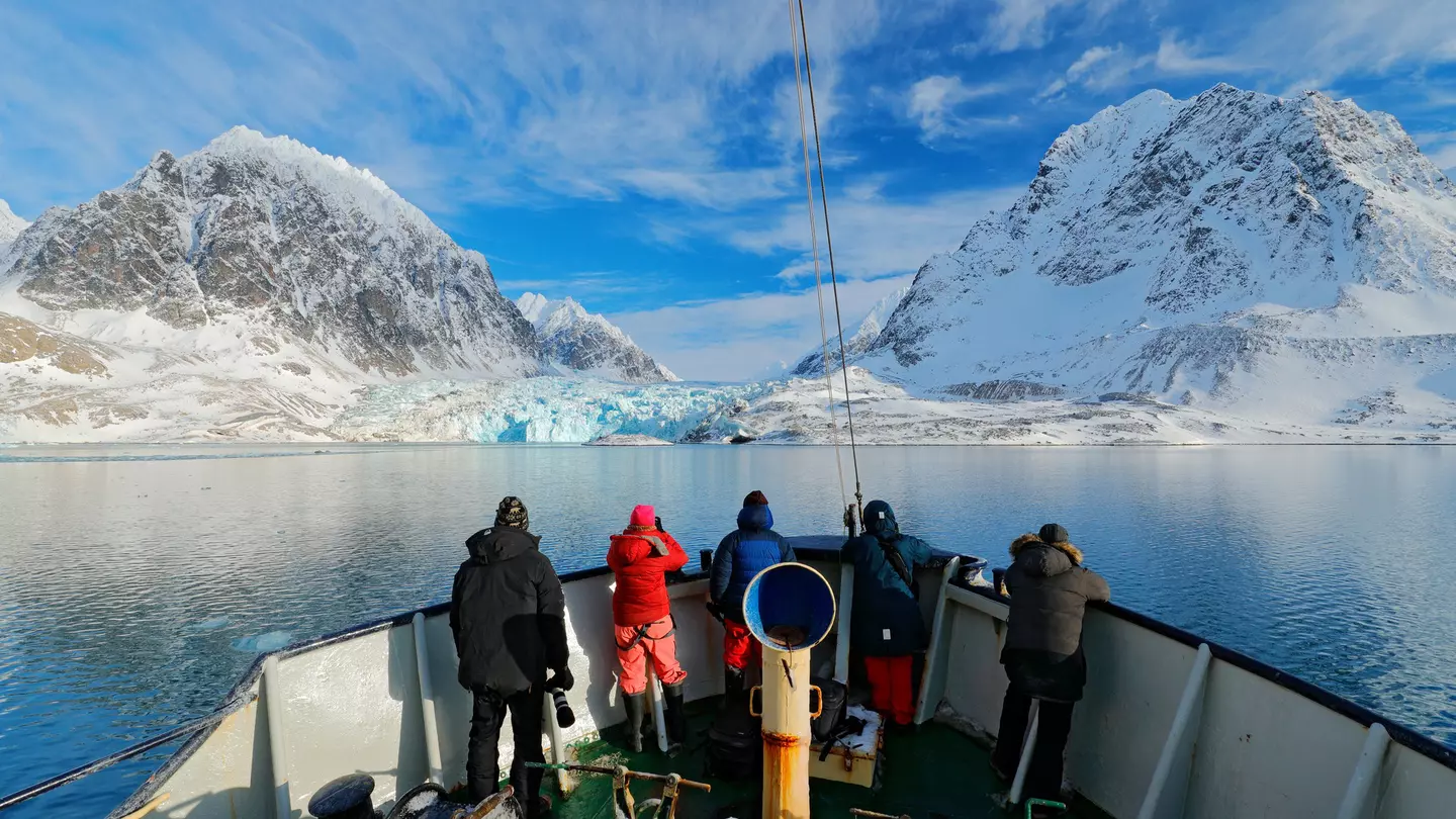 Four people at the front of a boat in blue water heading toward a blue glacier next to snow-covered mountains on shore.