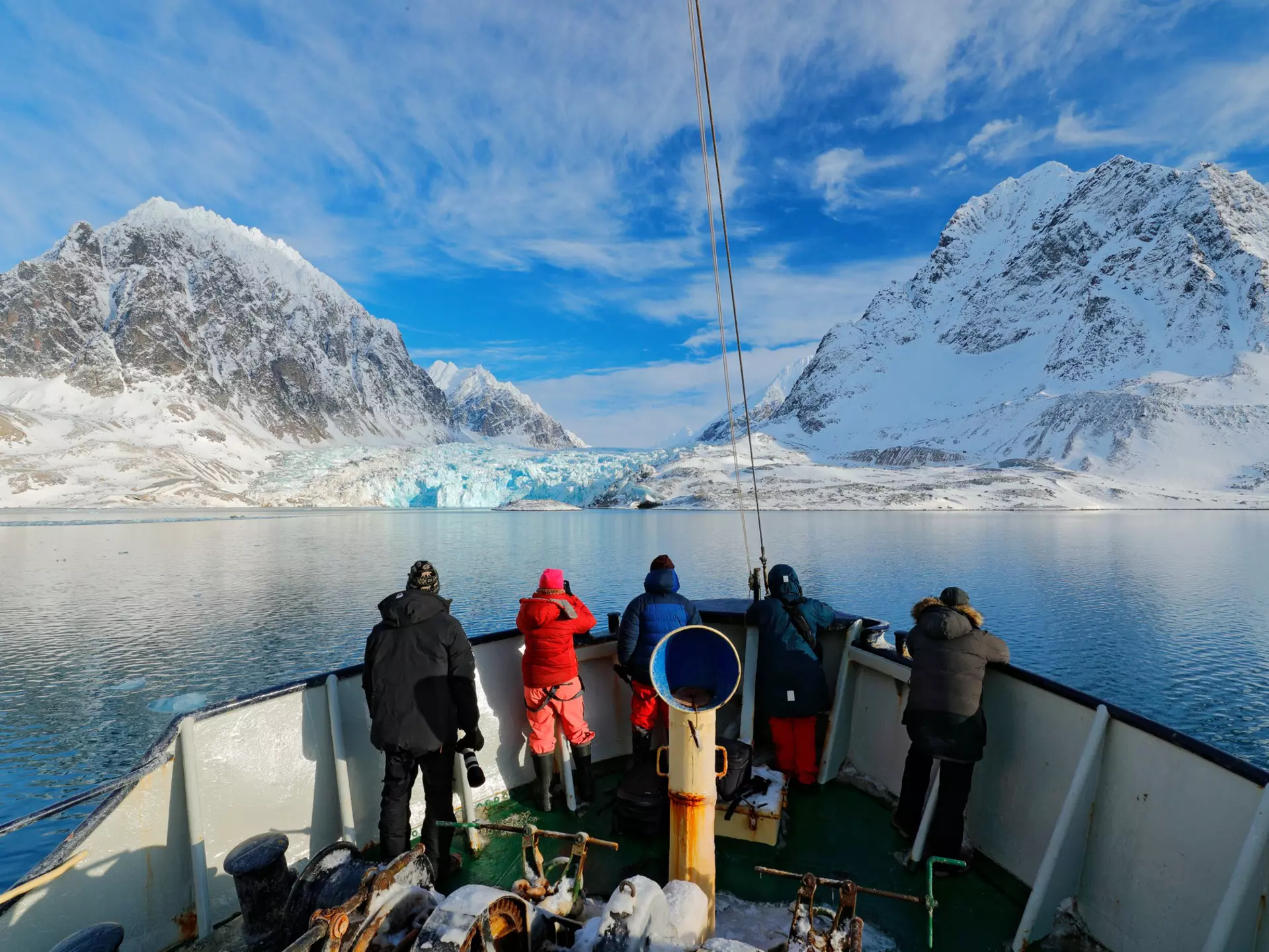 Four people at the front of a boat in blue water heading toward a blue glacier next to snow-covered mountains on shore.