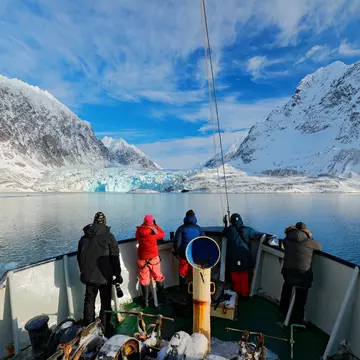 Four people at the front of a boat in blue water heading toward a blue glacier next to snow-covered mountains on shore.
