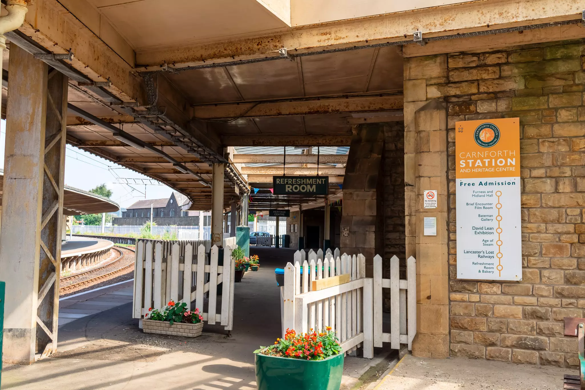 A white fence on a train station platform. Signs show the way to the Refreshment Room and Heritage Centre.