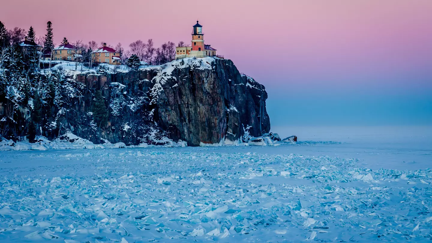 A lighthouse on the top of a rocky cliff glows pink the low sunlight. The lake below is frosty