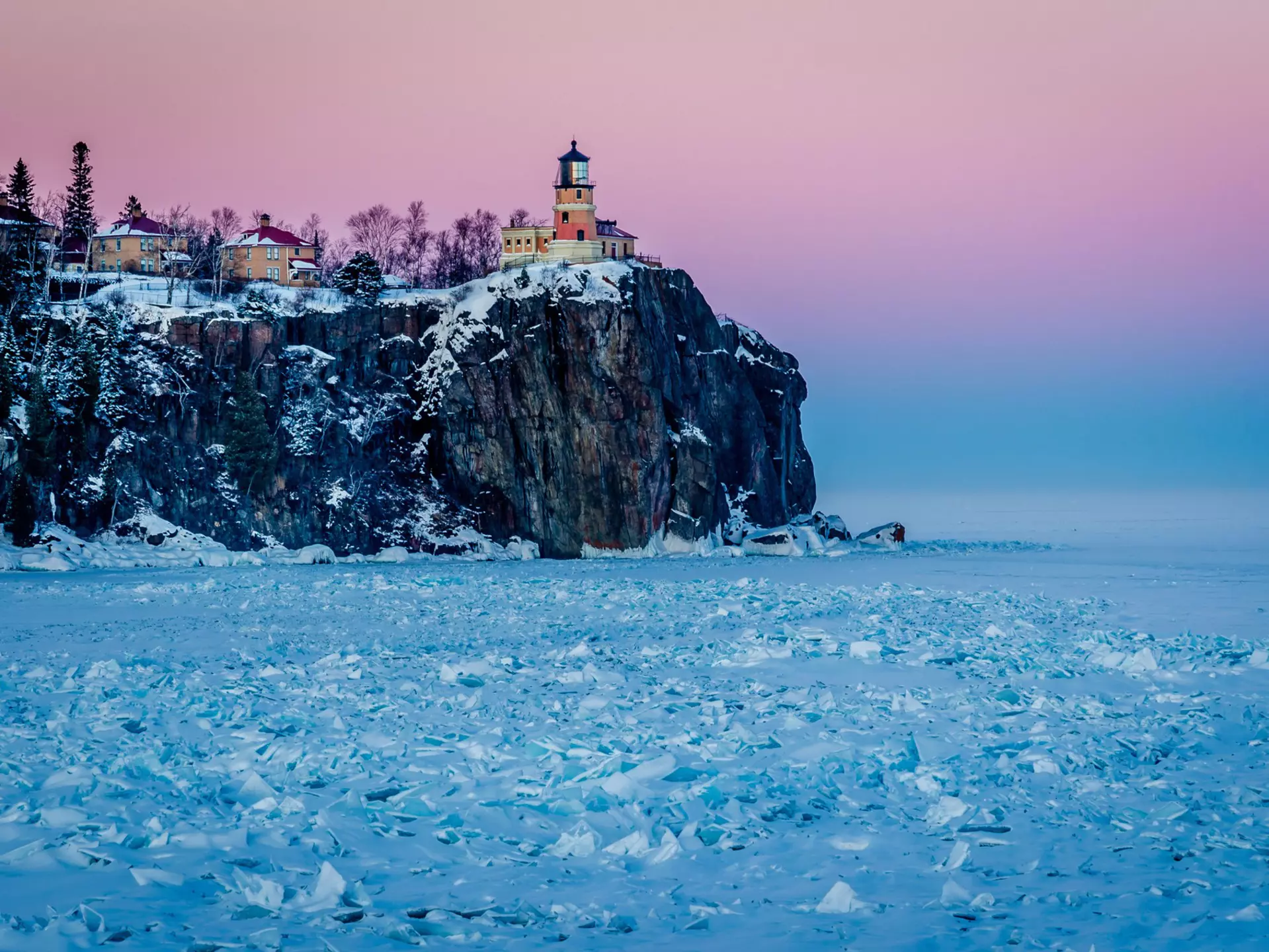 A lighthouse on the top of a rocky cliff glows pink the low sunlight. The lake below is frosty