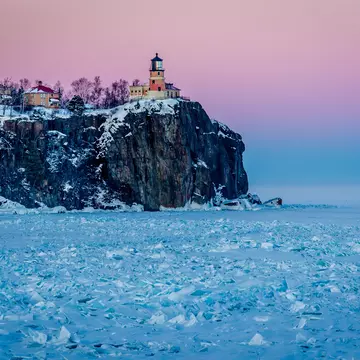 A lighthouse on the top of a rocky cliff glows pink the low sunlight. The lake below is frosty