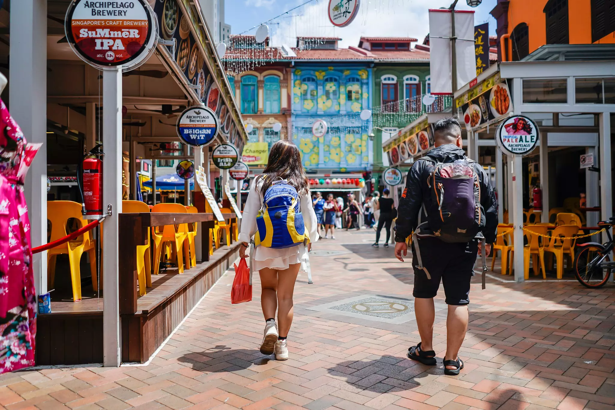two people in backpacks walk down a brick street lined with restaurants with yellow chairs and shops