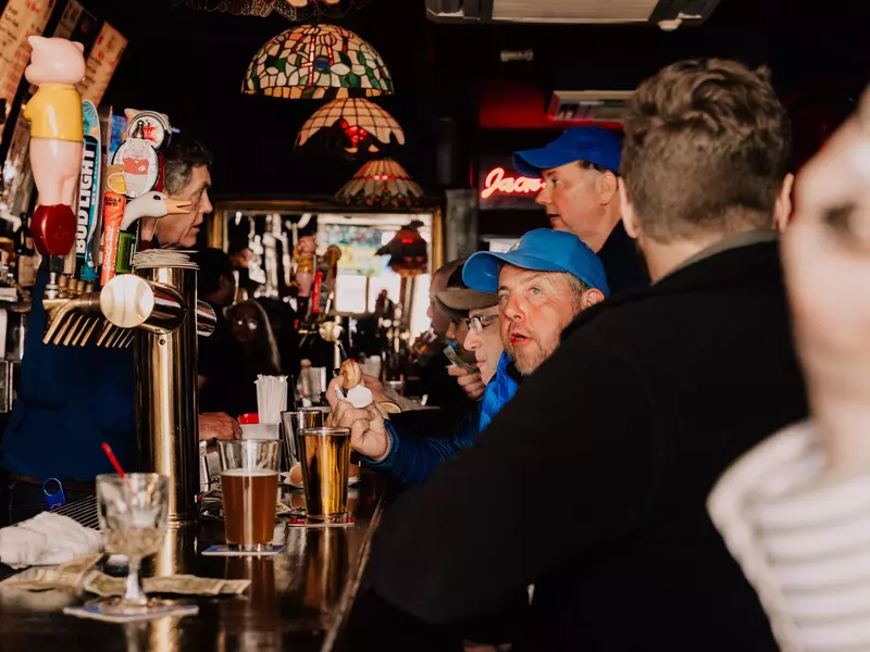 A man leans over the taps at a bar to talk with the patrons, who are all men, wearing hats and drinking pints of beer