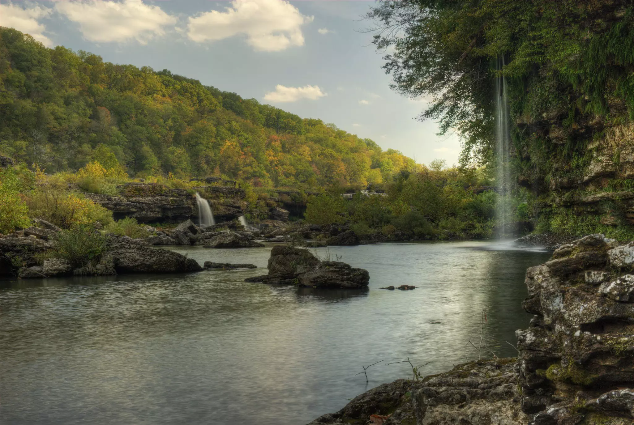A thin, high waterfall to the right and a lower, denser waterfall in the left distance with a small body of water in front. In the far distance is a forested hillside with just-yellowing leaves.
