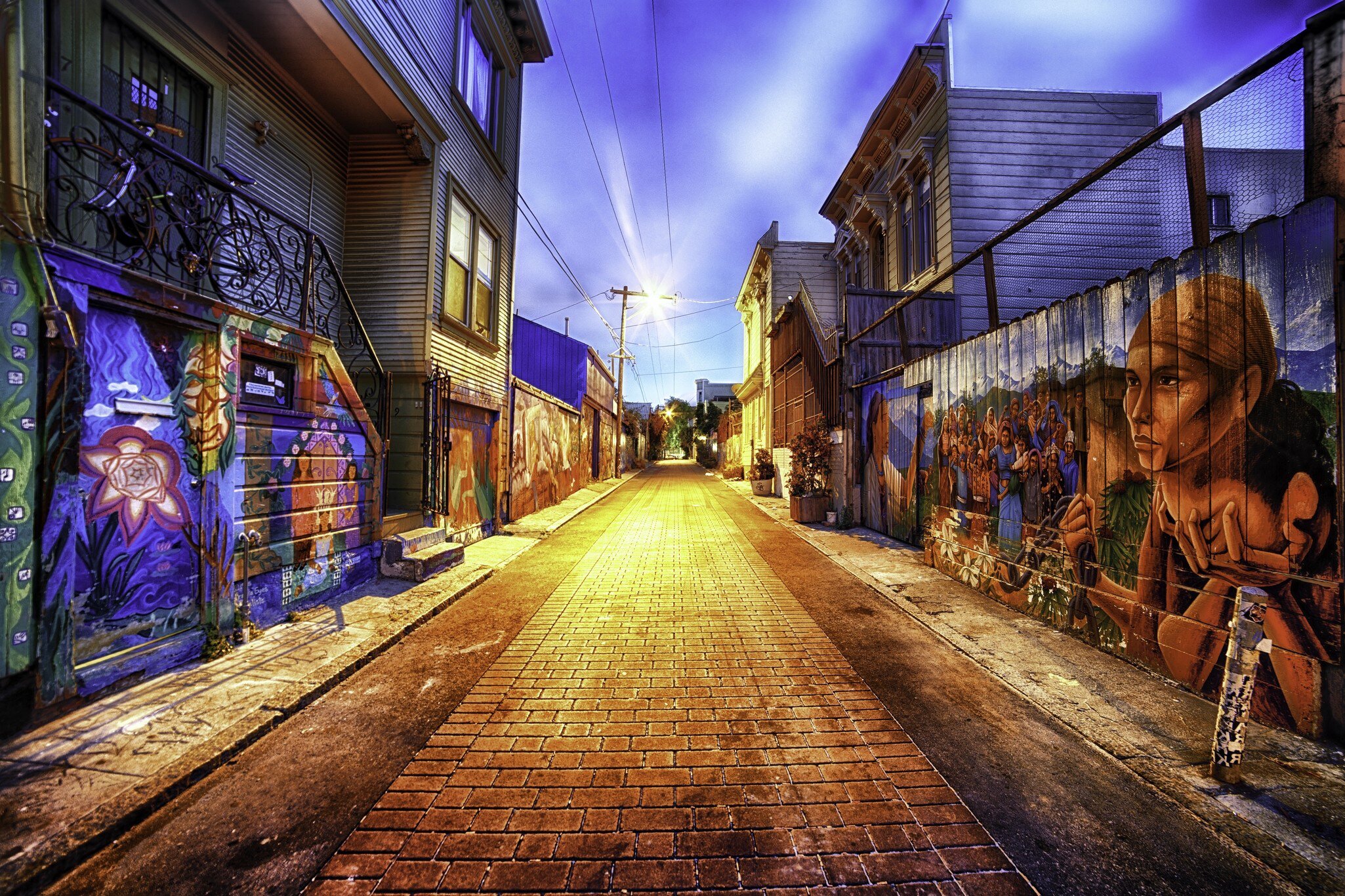 Painted figures and flowers cover both walls of a brick-paved lane at dusk.