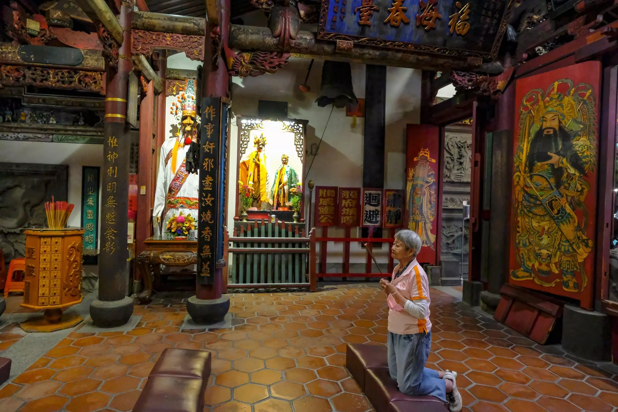 A woman kneels in prayer at a temple decorated with inscriptions in Chinese characters and depictions of deities.