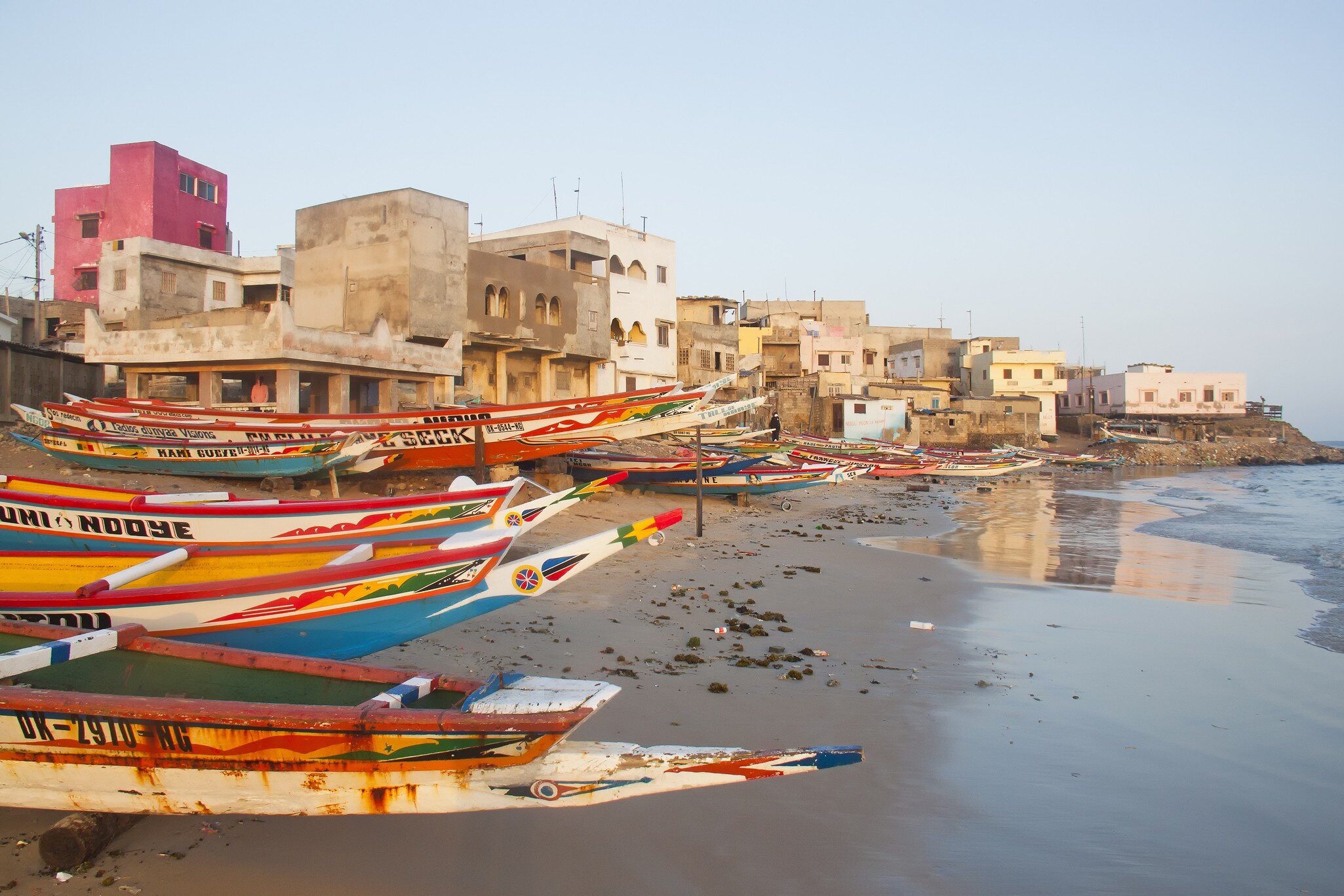 Colourful wooden fishing boats line a sandy shore below pink and white concrete houses, with calm water to the right