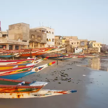 Colourful wooden fishing boats line a sandy shore below pink and white concrete houses, with calm water to the right