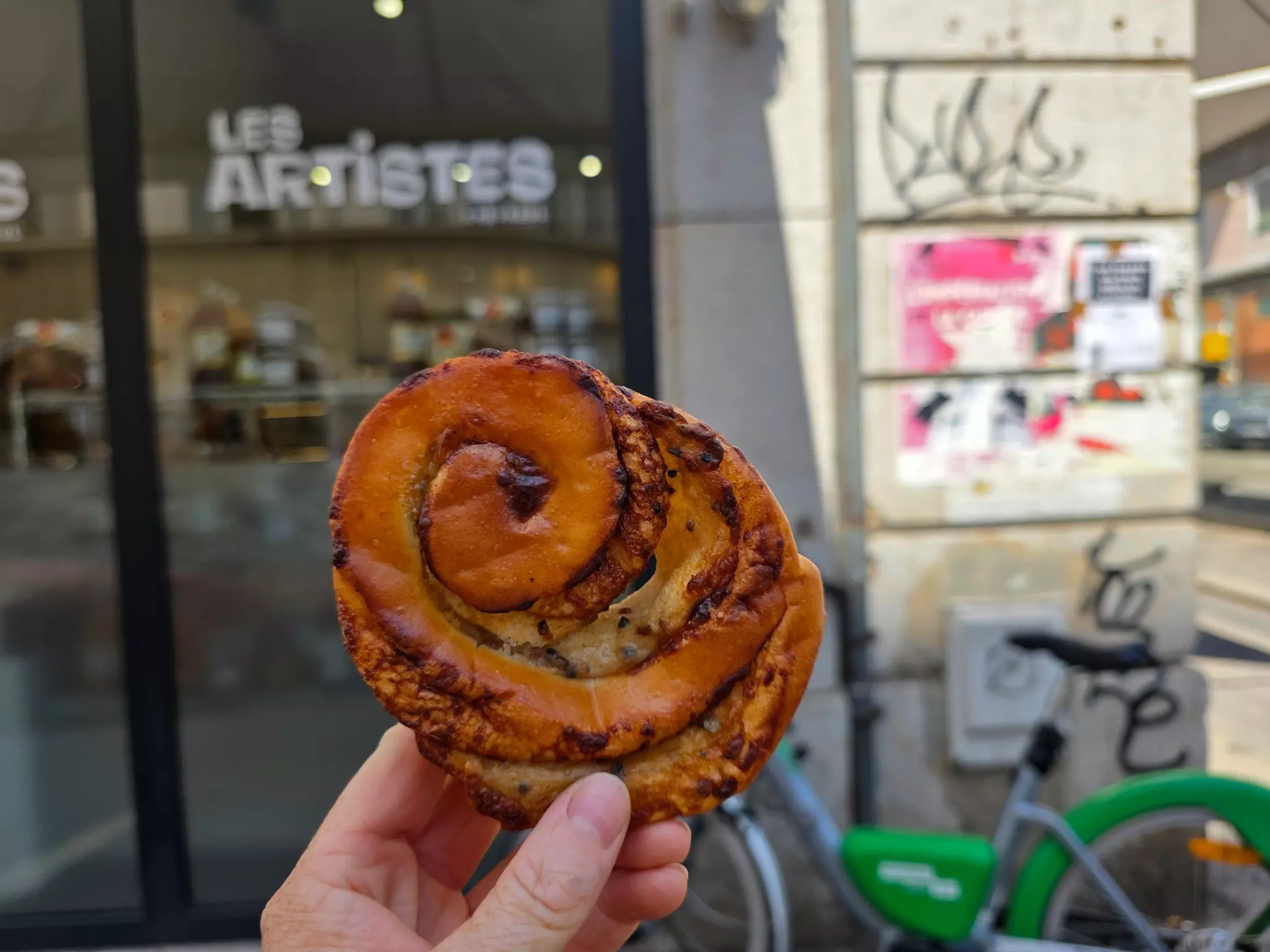A hand holds up a fresh pastry swirl outside a bakery