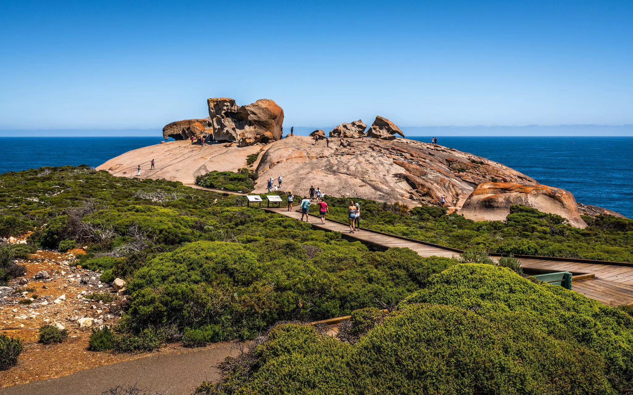 Visitors walk on a wooden boardwalk towards distinctive rock formations at the edge of a cliff.