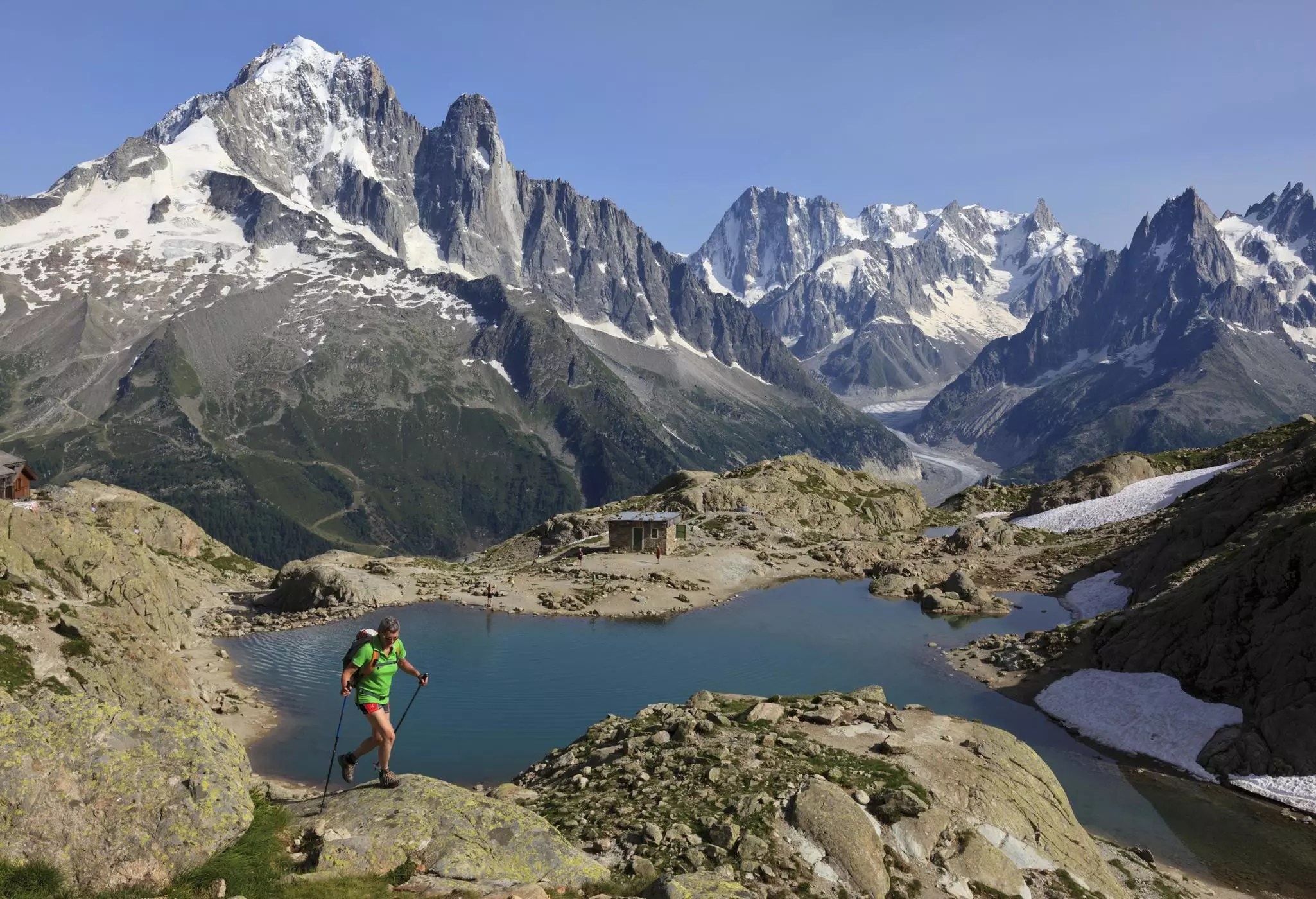 A hiker crosses rocky terrain in mountains high above an alpine lake with a small mountain hut on the shores.